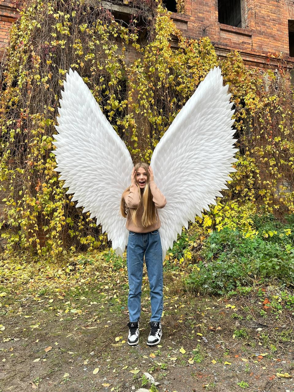 May include: A person wearing a brown sweater and blue jeans is standing in front of a brick wall covered in vines. They are holding large white angel wings.
