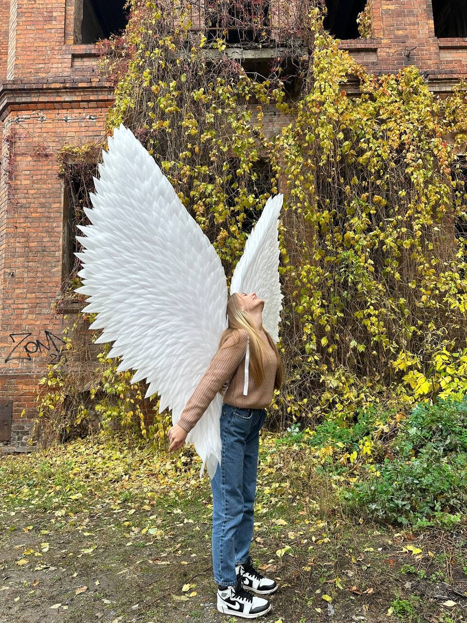 May include: A person wearing a brown sweater and blue jeans with large white angel wings. The person is standing in front of a brick building with vines growing on it.