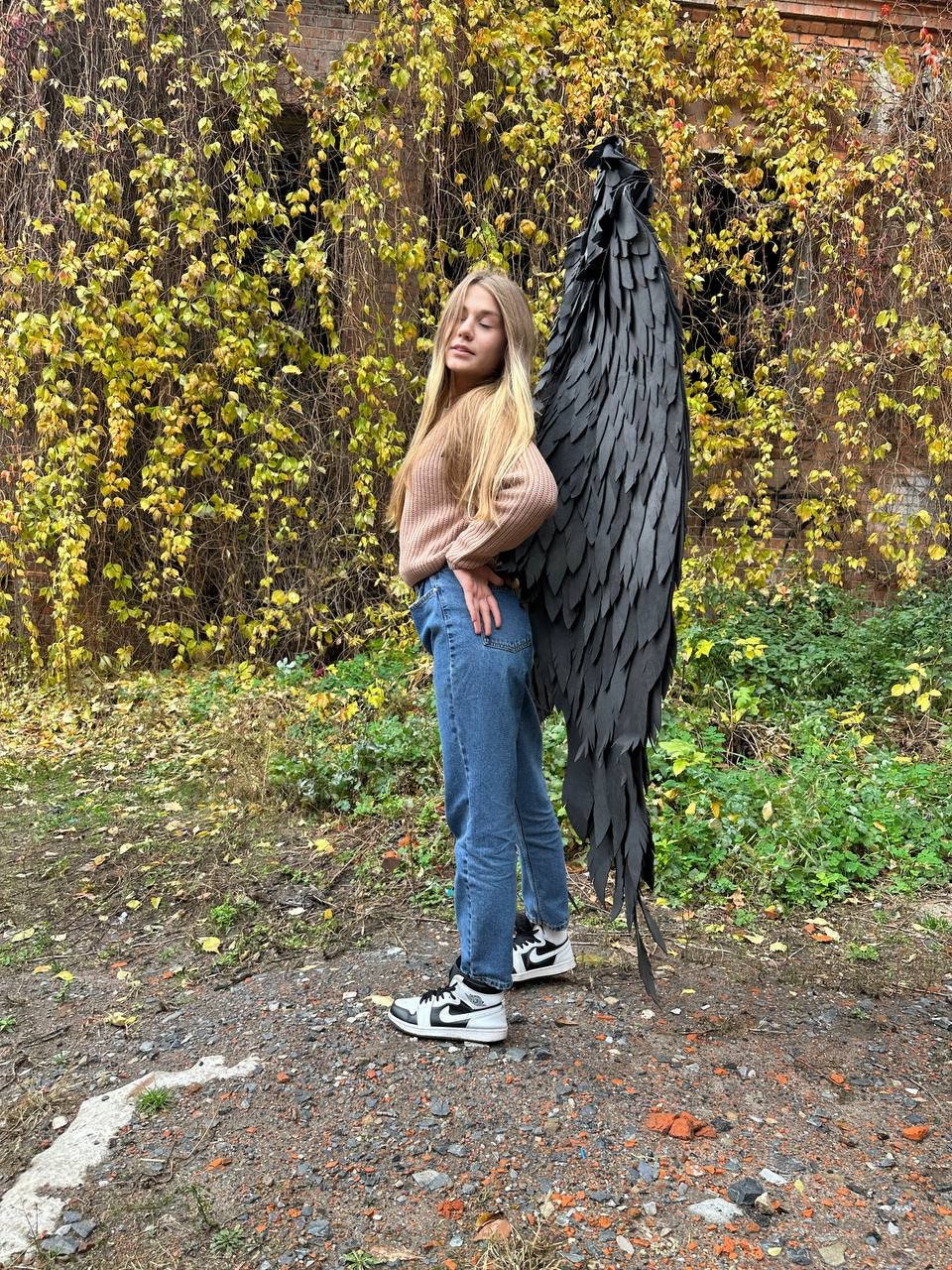 May include: A young person wearing blue jeans and a brown sweater stands in front of a wall of green vines. They are wearing black and white sneakers and holding a large black feathered wing behind them.