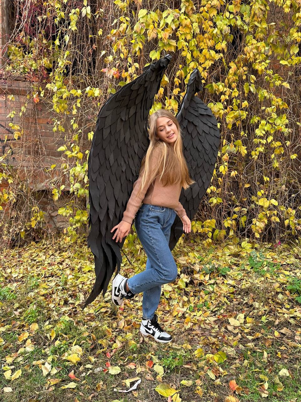 May include: A young person wearing blue jeans and a brown sweater poses with large black feathered wings. The wings are made of paper and have a realistic look. The person is standing in a grassy area with fallen leaves.