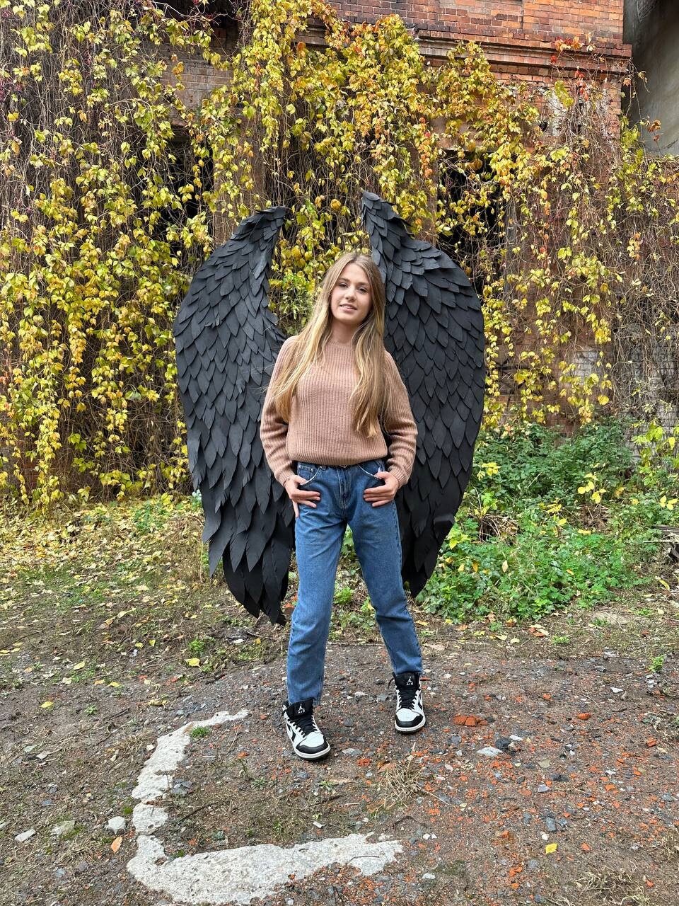 May include: A person wearing jeans and a brown sweater stands in front of a brick wall covered in vines. They are holding large black angel wings.