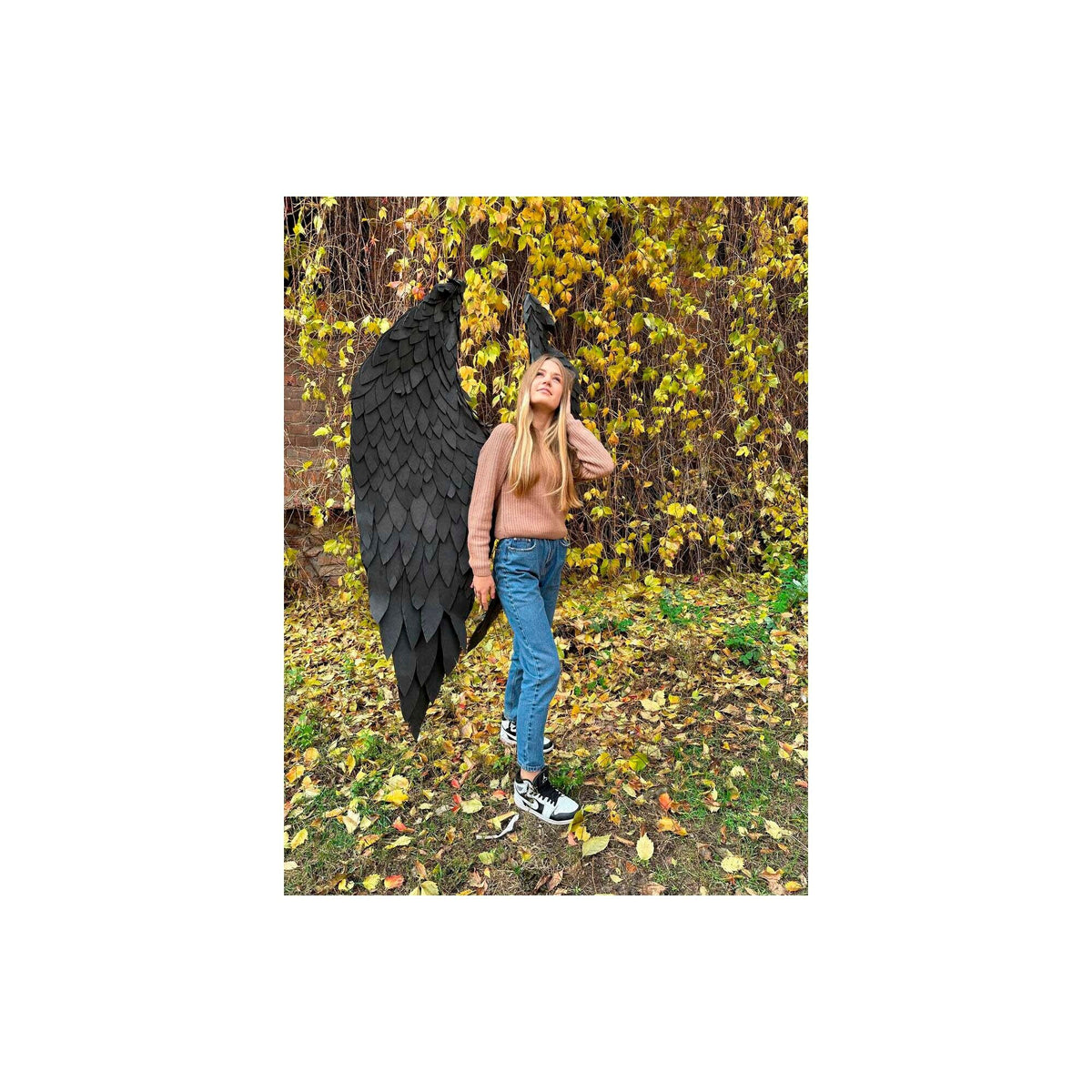 May include: A young person wearing blue jeans and a brown sweater stands in a forest setting with a large pair of black feathered wings behind them.