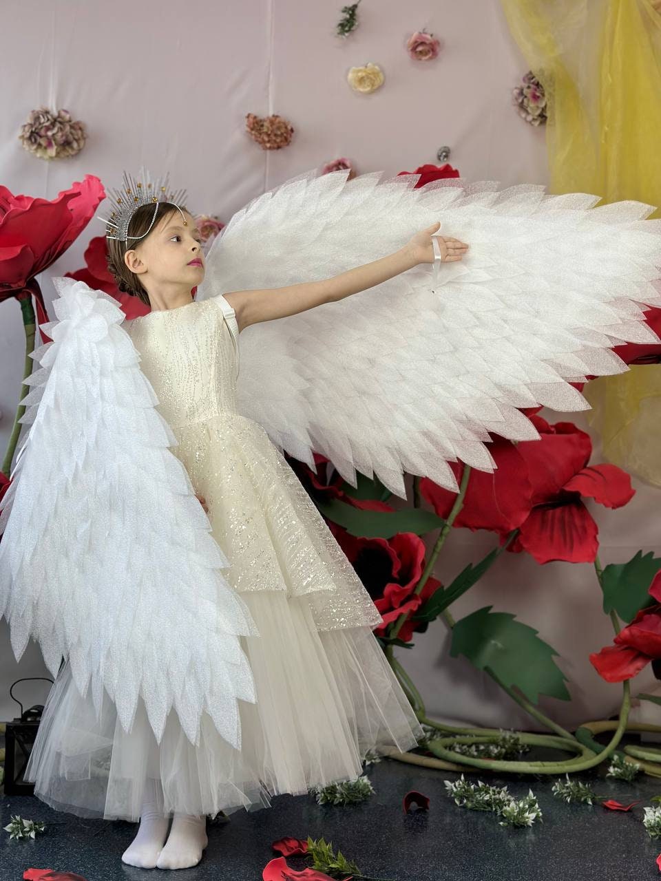 May include: A young person wearing a white dress and a silver crown, with large white angel wings, standing in front of a backdrop of red flowers.