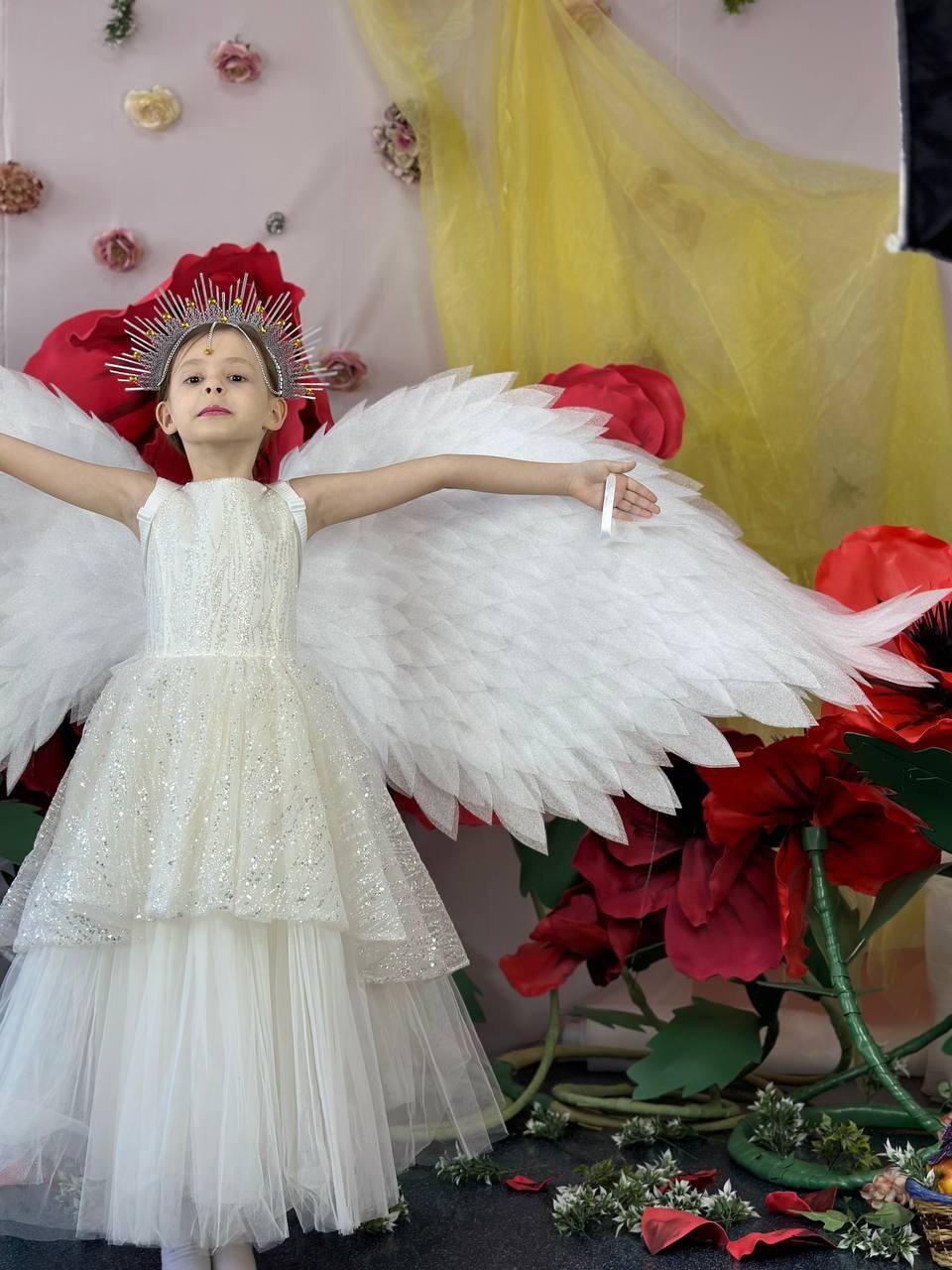 May include: A young person wearing a white dress with a sparkly bodice and large white angel wings. They are wearing a gold crown with a red jewel on top. The person is standing in front of a backdrop of red flowers and a yellow sheer curtain.