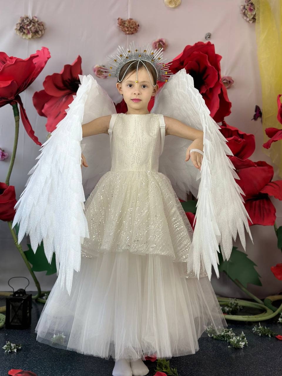 May include: A young girl wearing a white dress with a sparkly bodice and a white tulle skirt. She is wearing a silver crown and white angel wings. The background is a pink wall with red flowers.