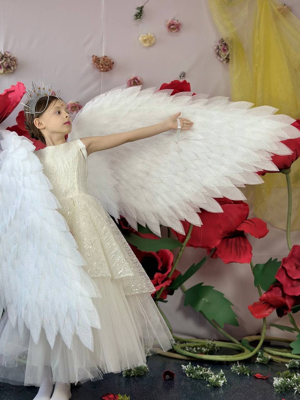May include: A young girl wearing a white dress and a silver crown, with large white feathered angel wings, standing in front of a backdrop of red flowers.