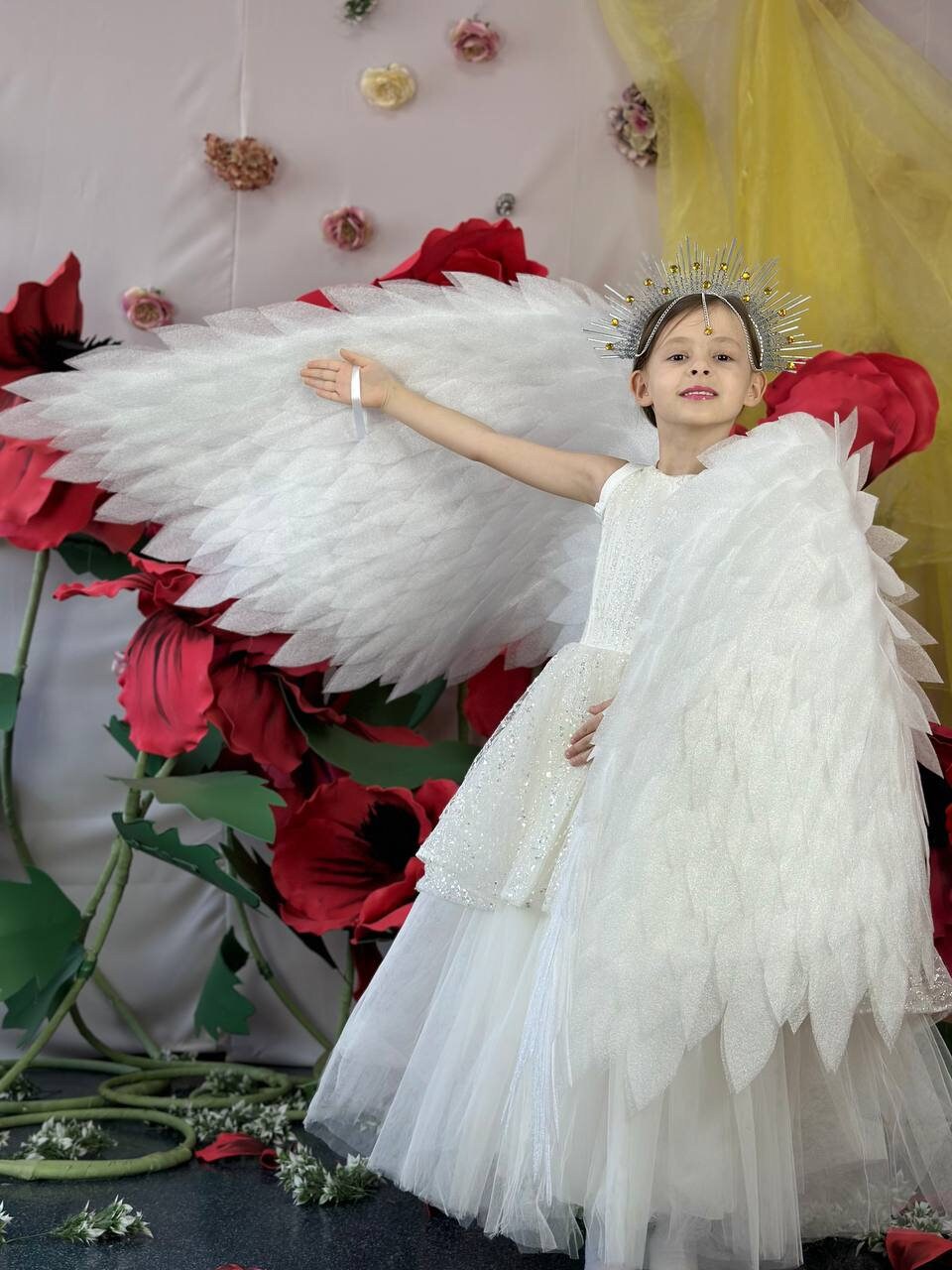 May include: A young girl wearing a white dress and angel wings with a silver crown on her head. She is standing in front of a backdrop of large red paper flowers.