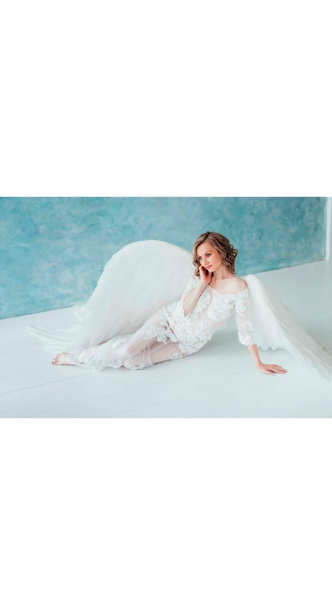May include: A woman wearing a white lace dress with sheer fabric and angel wings, posing on a white surface against a blue background.