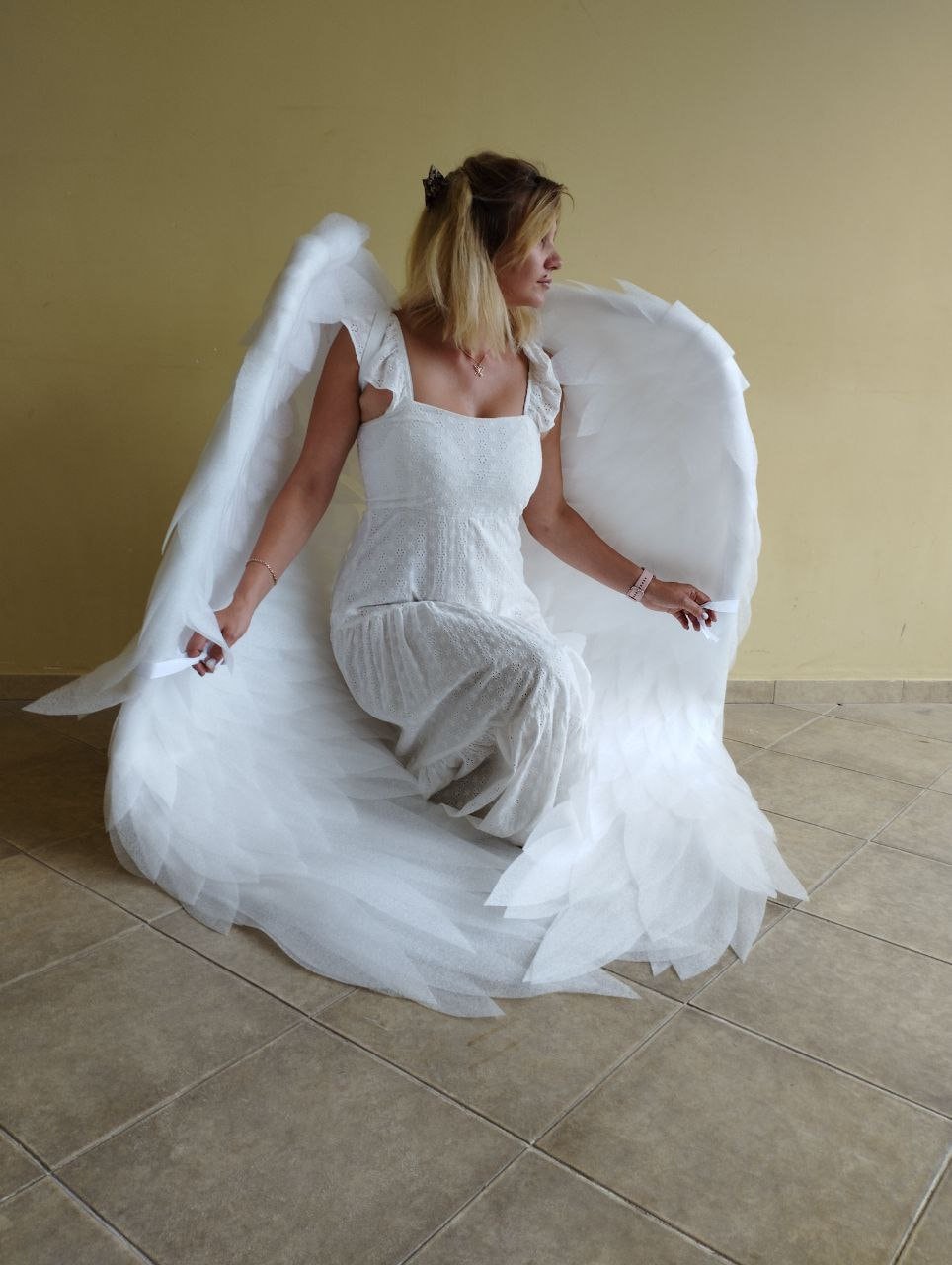 May include: A woman wearing a white dress with a white angel wing costume. The wings are large and feathery, and the woman is sitting on the floor with her legs crossed.