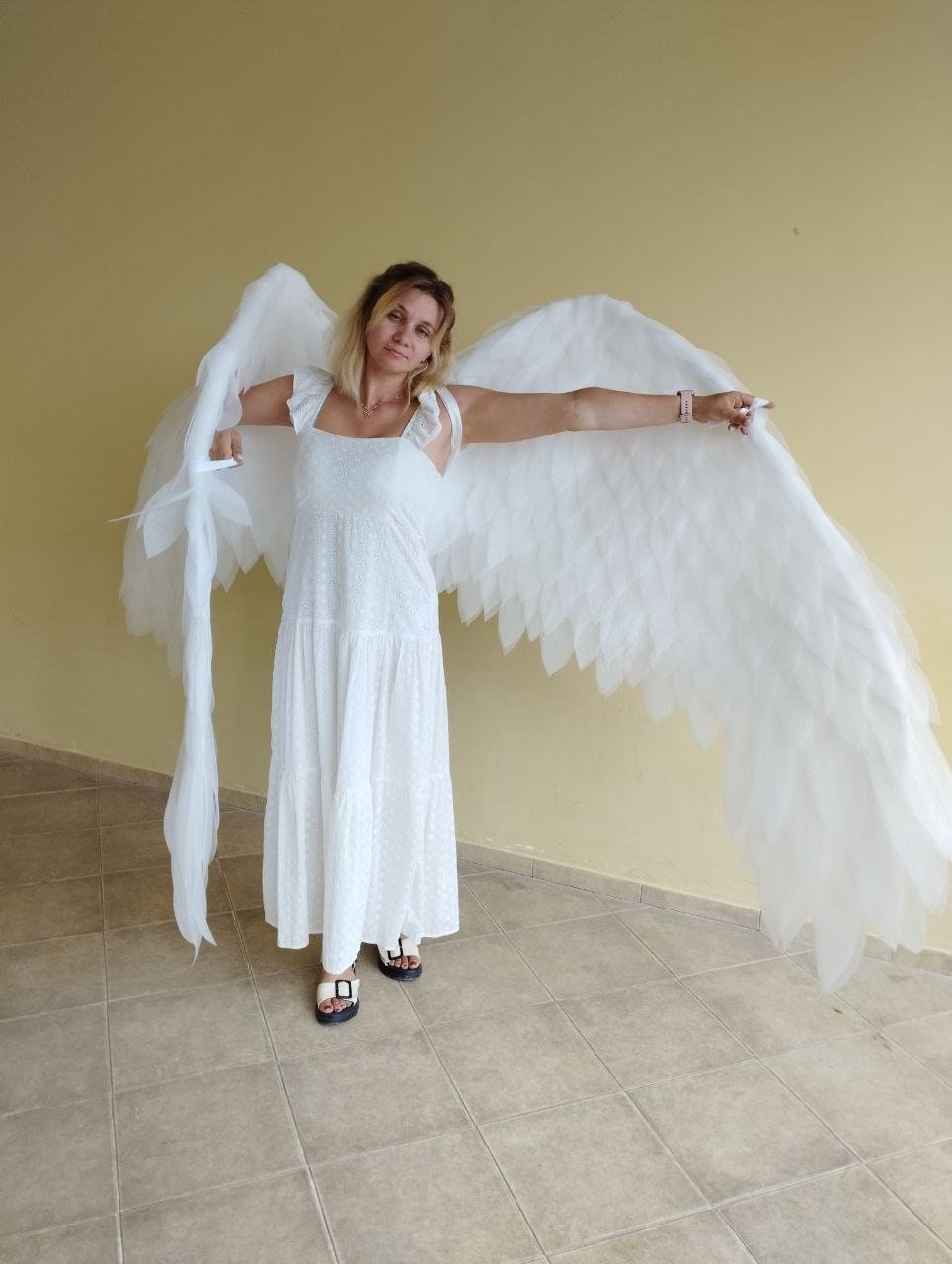 May include: A person wearing a white dress and large white angel wings. The wings are made of feathers and are spread out behind the person.