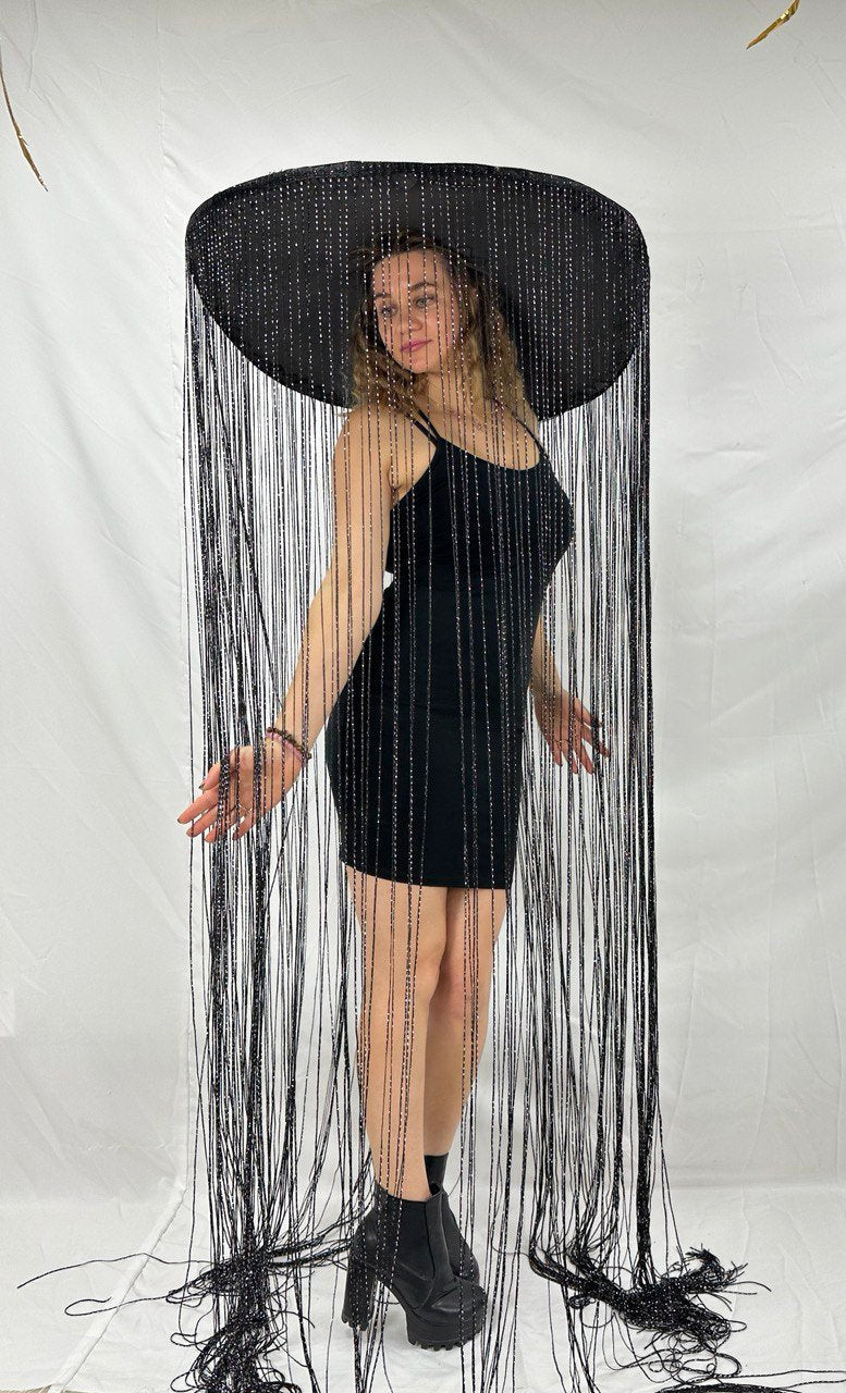 May include: A woman wearing a black dress and a large black hat with long fringe. The hat is wide-brimmed and the fringe hangs down to her ankles.