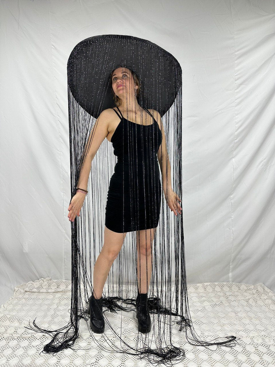 May include: A black wide-brimmed hat with long black fringe hanging from the brim. The hat is worn by a person wearing a black dress.