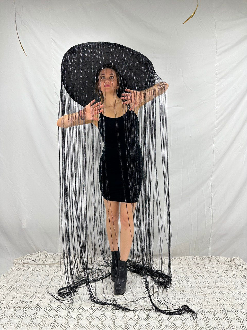 May include: A woman wearing a black velvet dress and a large black hat with long black strings hanging down. The hat is wide brimmed and the strings are beaded.