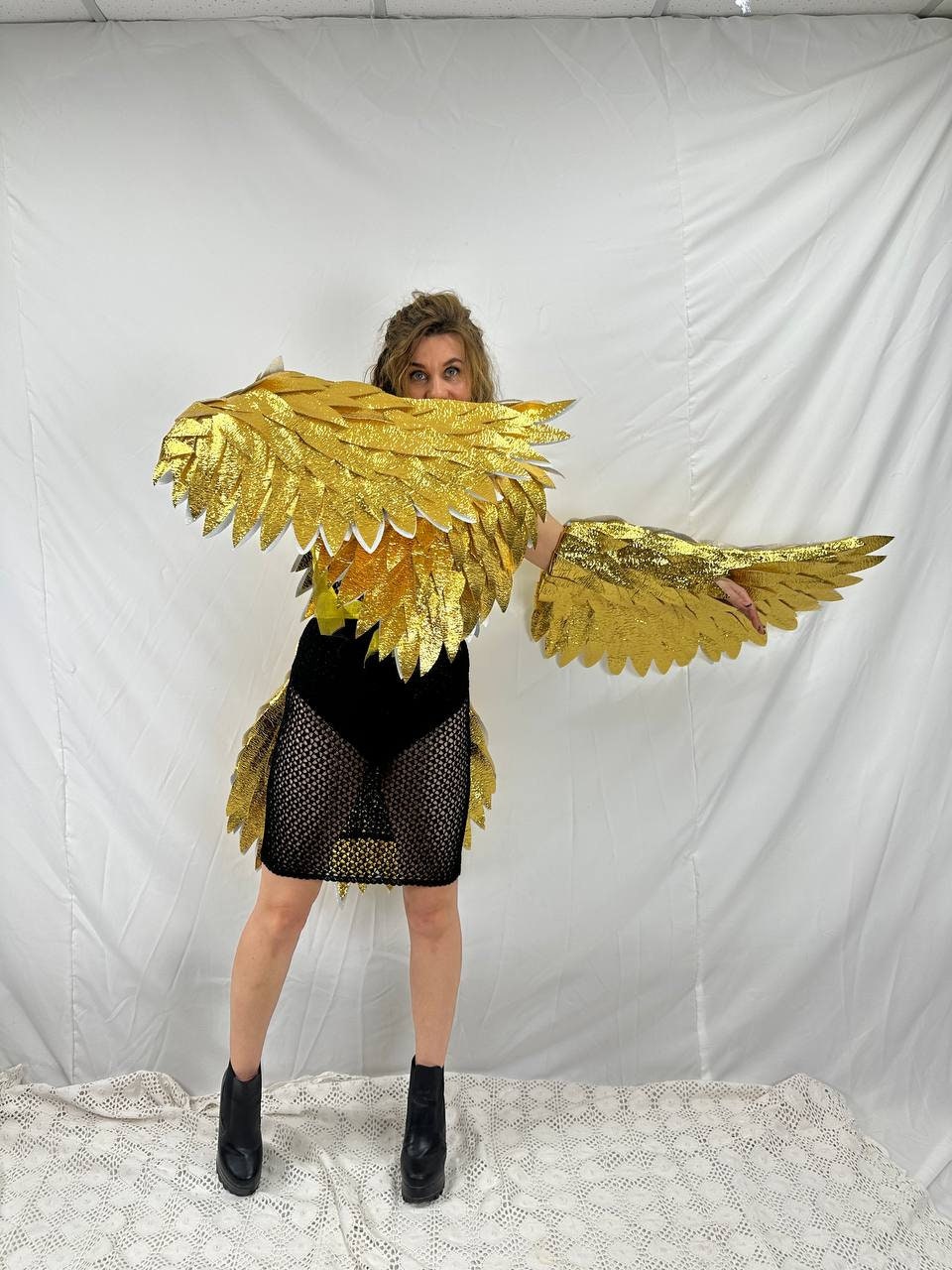 May include: A person wearing a gold-colored eagle wing costume. The costume includes large, layered wings and arm pieces. The person is also wearing a black dress and black boots. The background is a white sheet.