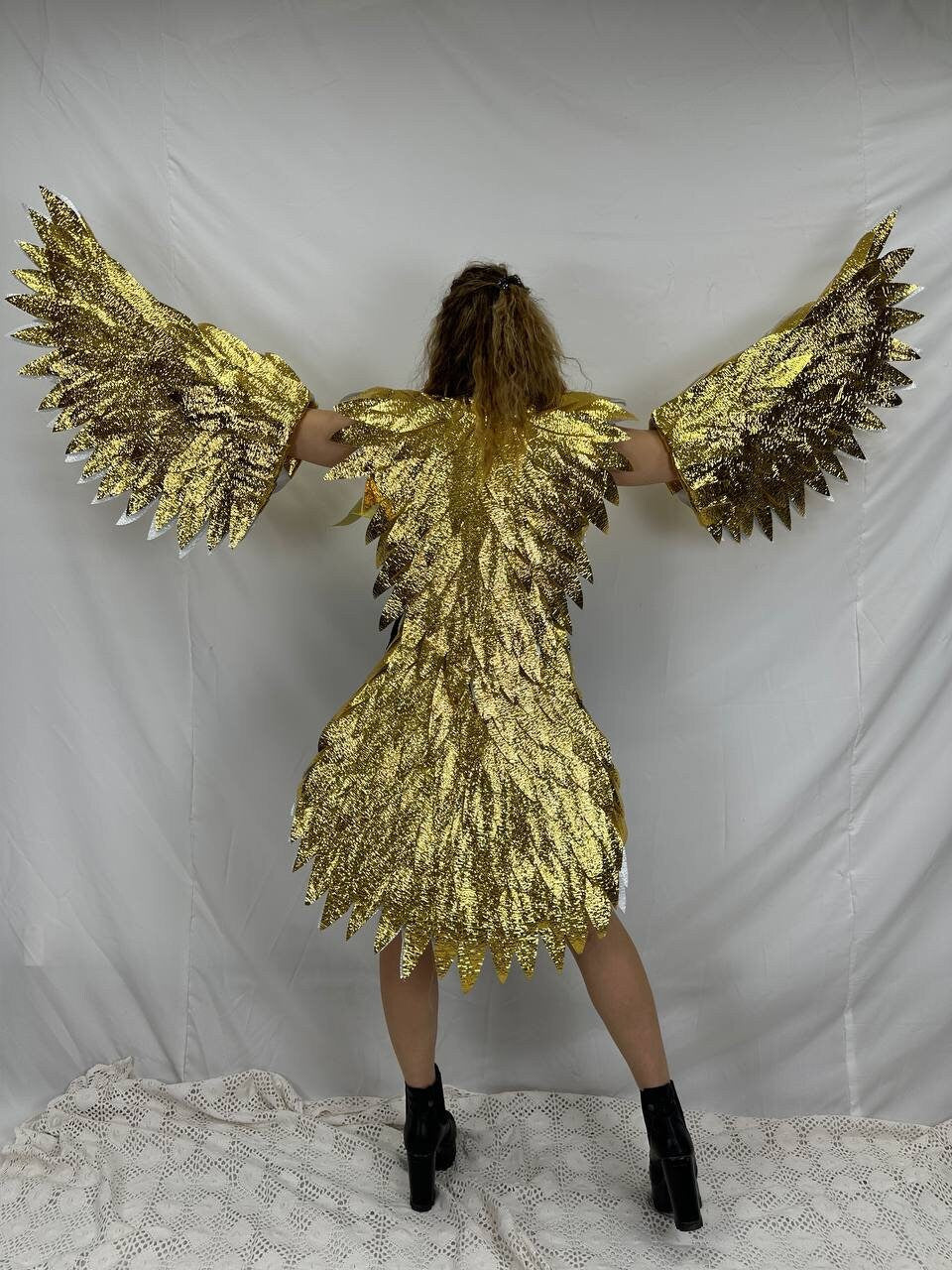 May include: A golden eagle costume with large, detailed wings and a matching bodice. The wings are outstretched, showcasing the intricate feather design and shimmering gold material. The costume is worn with black boots.