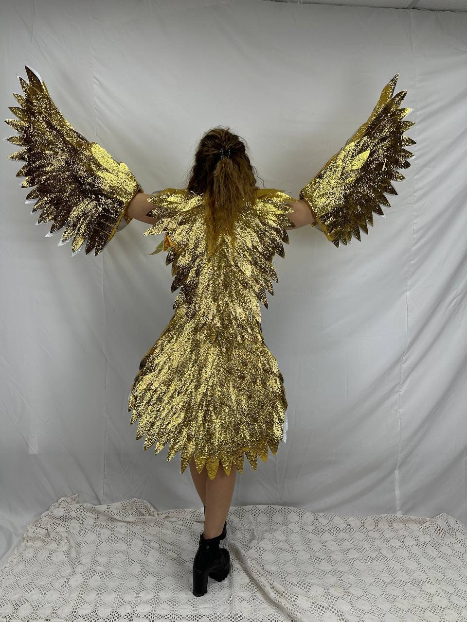 May include: A person wearing a gold costume with large wings. The costume is made of shiny, metallic material and has a feathered design. The person is standing on a white surface.