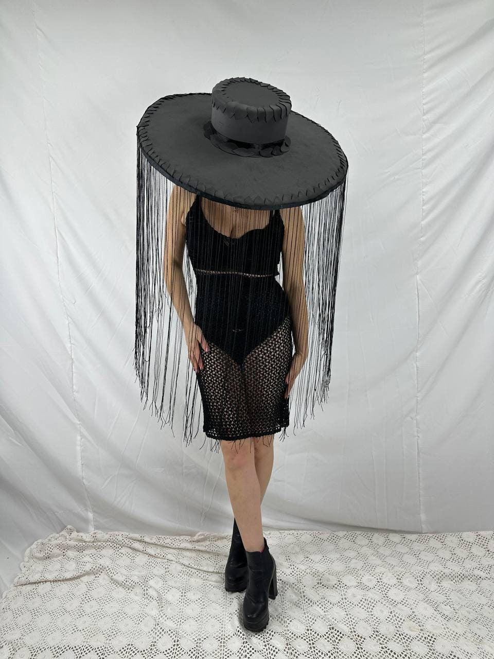 May include: A black wide-brimmed hat with fringe hanging from the brim. The hat is worn by a person wearing a black mesh dress and black boots.