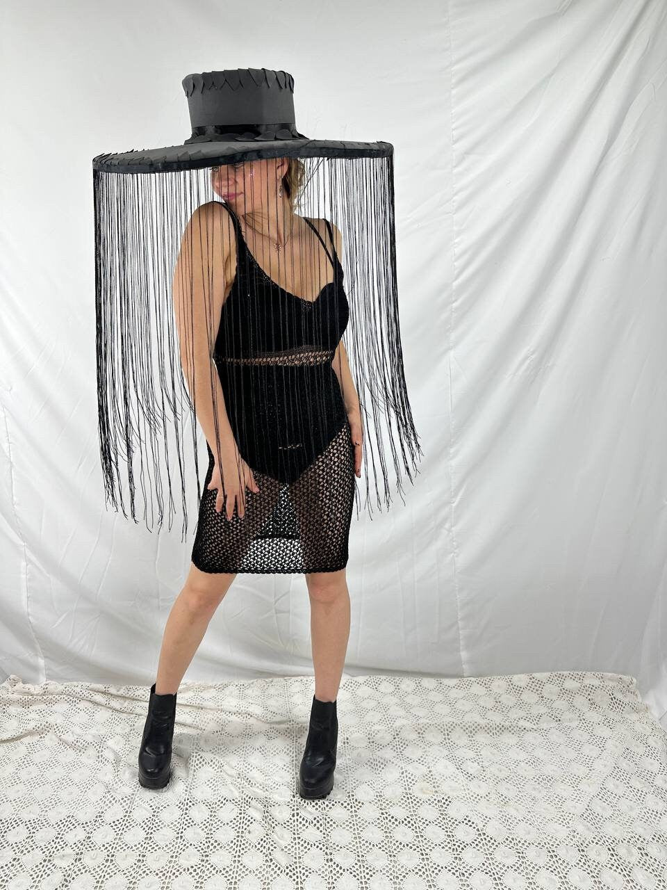 May include: A black wide-brimmed hat with fringe hanging from the brim. The hat is worn by a person wearing a black mesh dress and black boots.