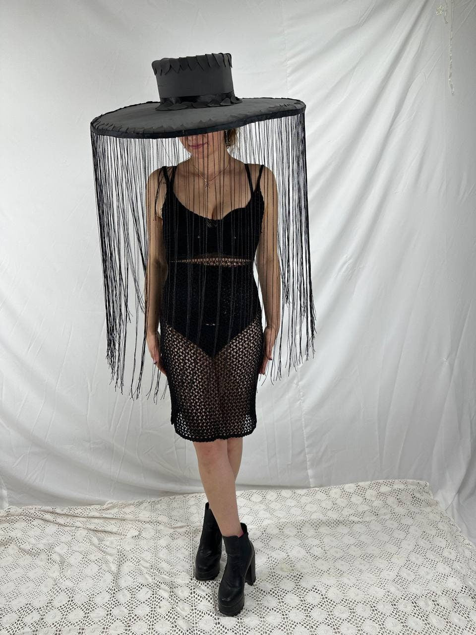 May include: A black wide-brimmed hat with fringe hanging from the brim. The hat is worn by a person wearing a black mesh dress and black boots.