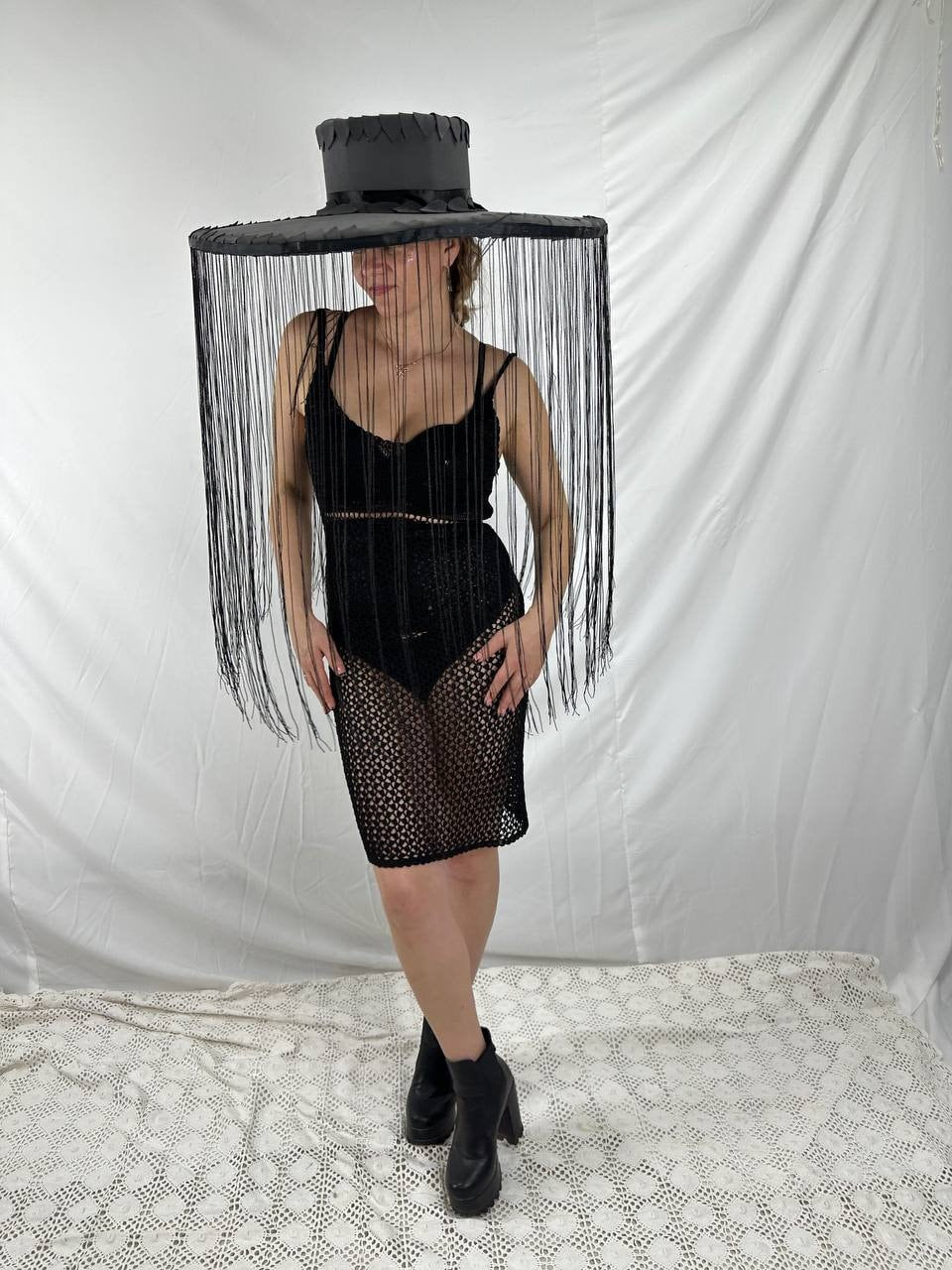 May include: A black wide-brimmed hat with a fringe trim. The hat is worn by a person wearing a black mesh dress and black boots.