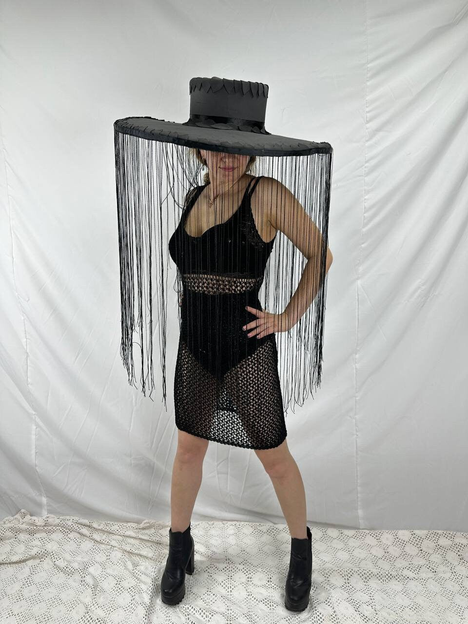 May include: A black wide-brimmed hat with fringe hanging from the brim. The hat is worn by a person wearing a black mesh dress and black boots.