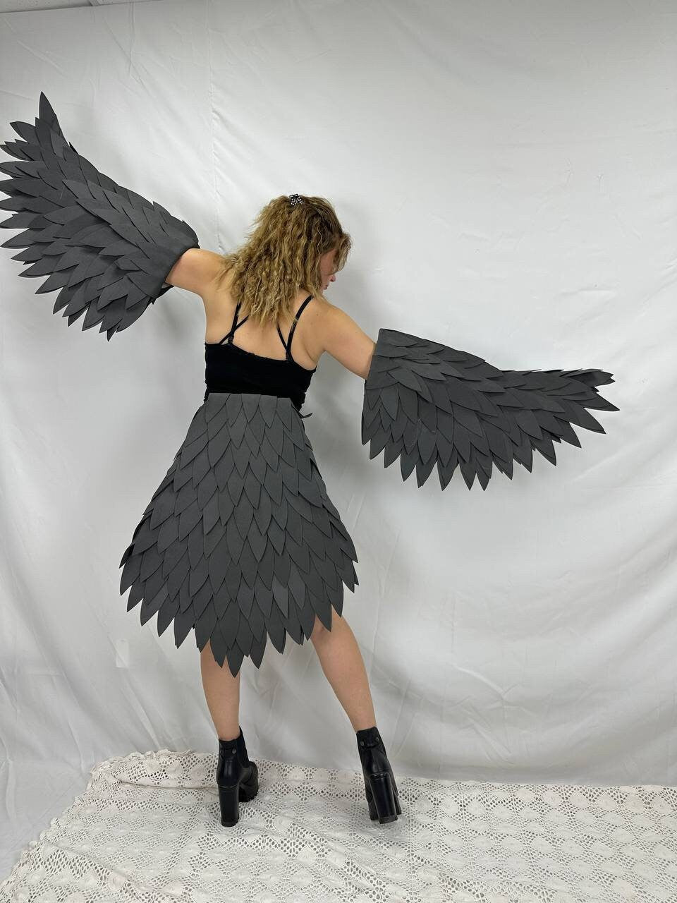 May include: A person wearing a black top and a skirt made of gray fabric resembling feathers. The person is wearing black boots and has their arms outstretched, holding gray feathered wings.