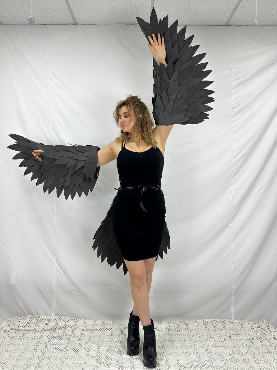 May include: A person wearing a black dress with black wings made of feathers. The wings are attached to the arms and the back of the dress.