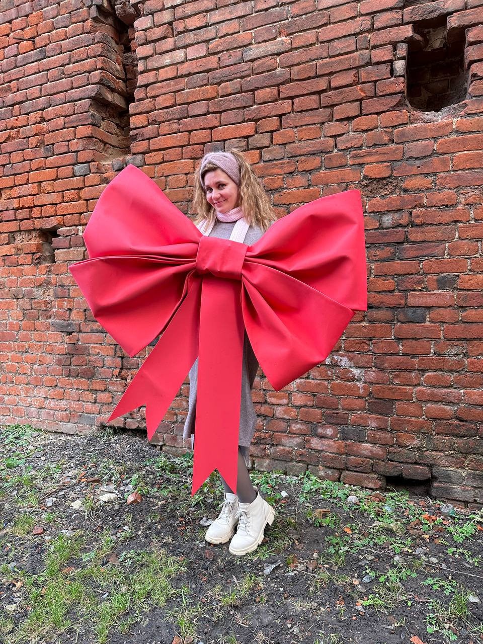 May include: A large, bright red bow with long streamers. The bow is held in front of a person wearing a gray dress and white sneakers. The background is a weathered brick wall.