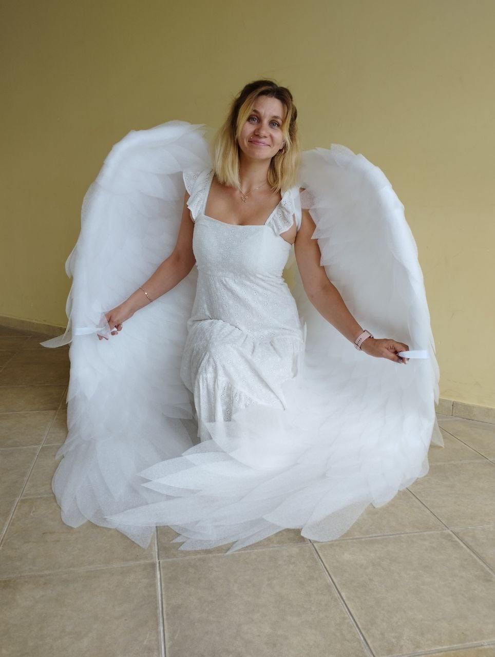May include: A woman wearing a white dress with a white angel wing costume. The wings are large and feathery, and the woman is smiling.