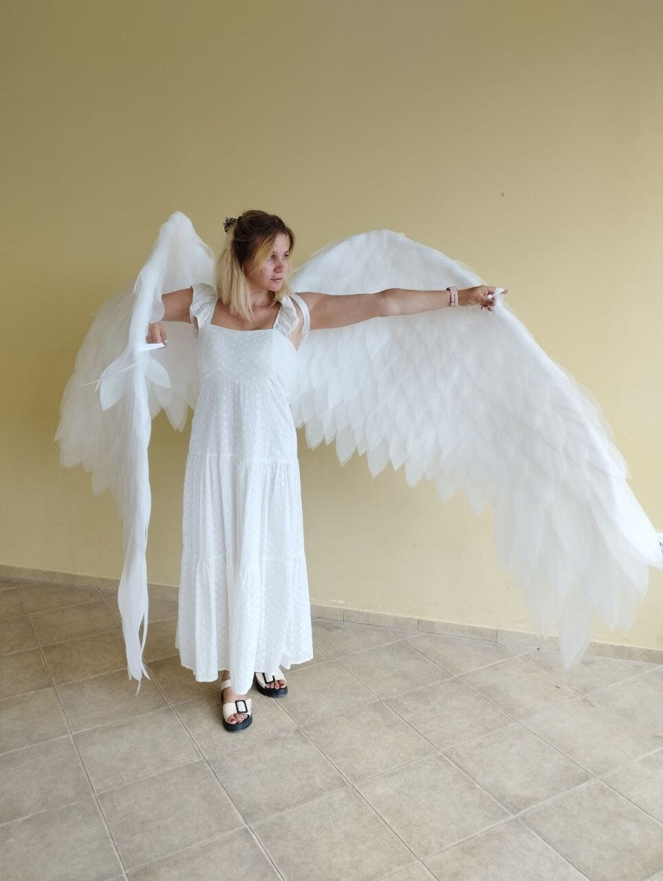 May include: A person wearing a white dress with white angel wings. The wings are large and feathery, and the person is standing in front of a light yellow wall.