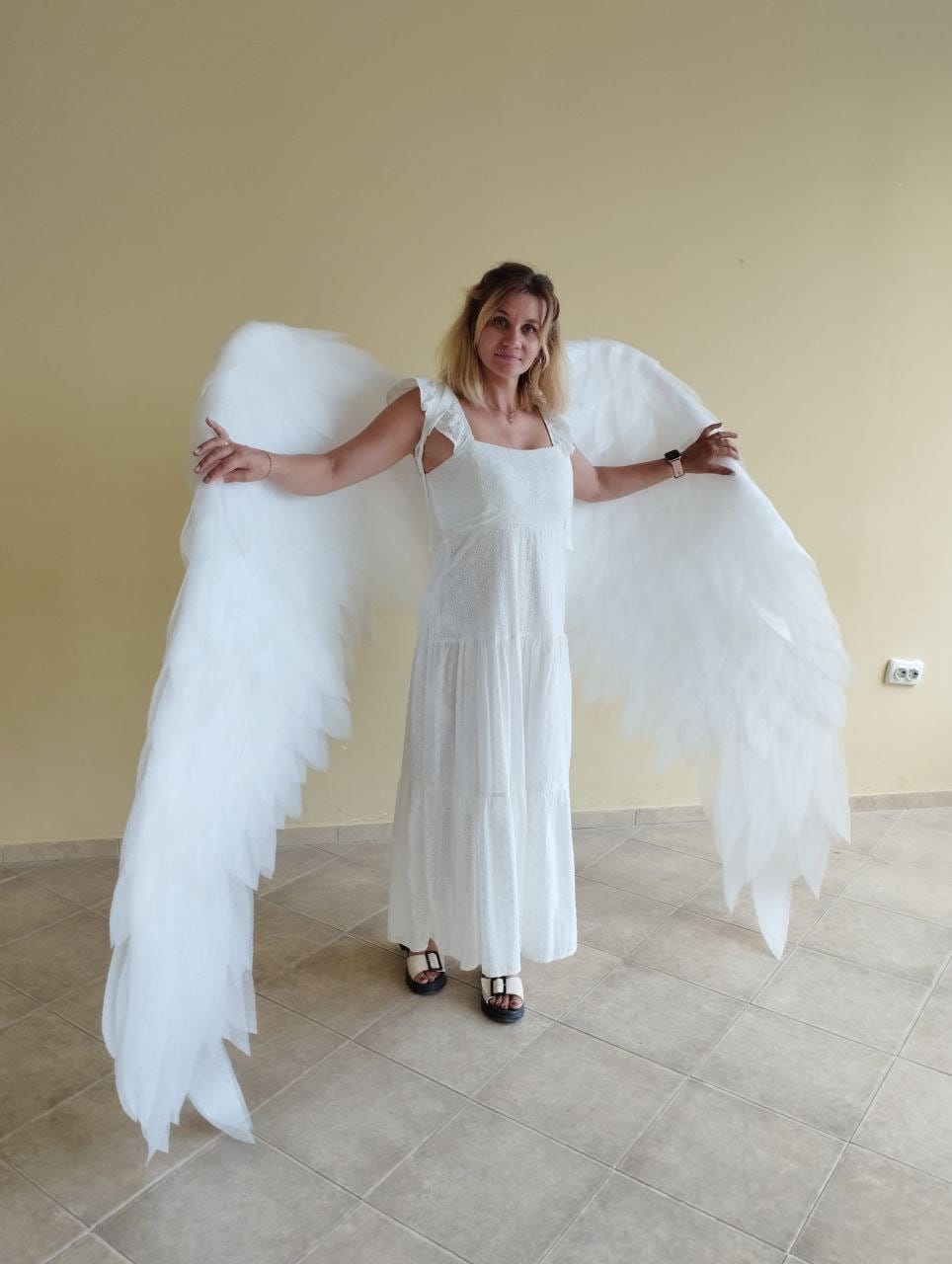 May include: A person wearing a white dress with a pair of white angel wings. The wings are large and feathery, and the person is standing in a room with tiled floors.
