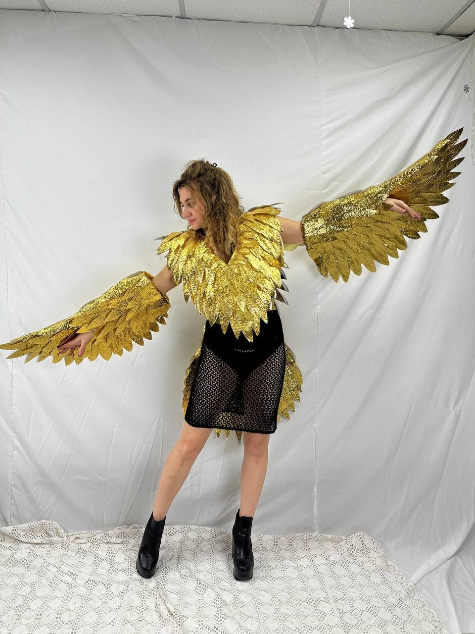 May include: A person wearing a gold sequined eagle costume with large, outstretched wings. The costume includes a black mesh dress and black boots. The wings and shoulder piece are made of gold sequins.