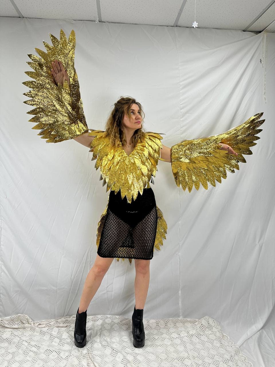 May include: A person wearing a gold-colored bird costume with large, outstretched wings. The costume includes a feathered collar and arm pieces, paired with a black skirt and platform boots. The wings are detailed with a textured, glittery finish.