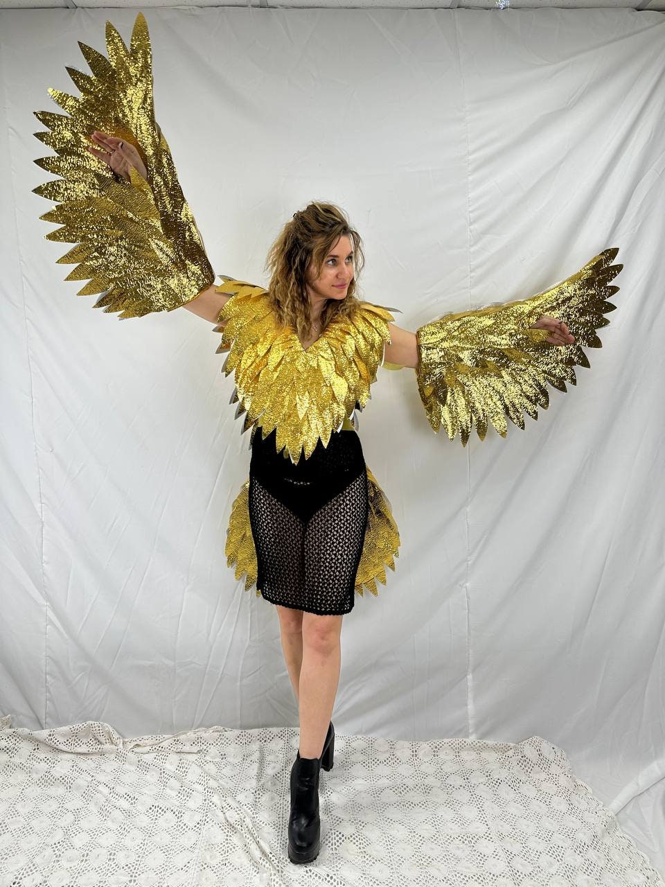 May include: A golden eagle costume with large, feathered wings and a matching bodice. The costume includes a black, open-weave skirt and black platform boots. The wings are outstretched, creating a dramatic pose.