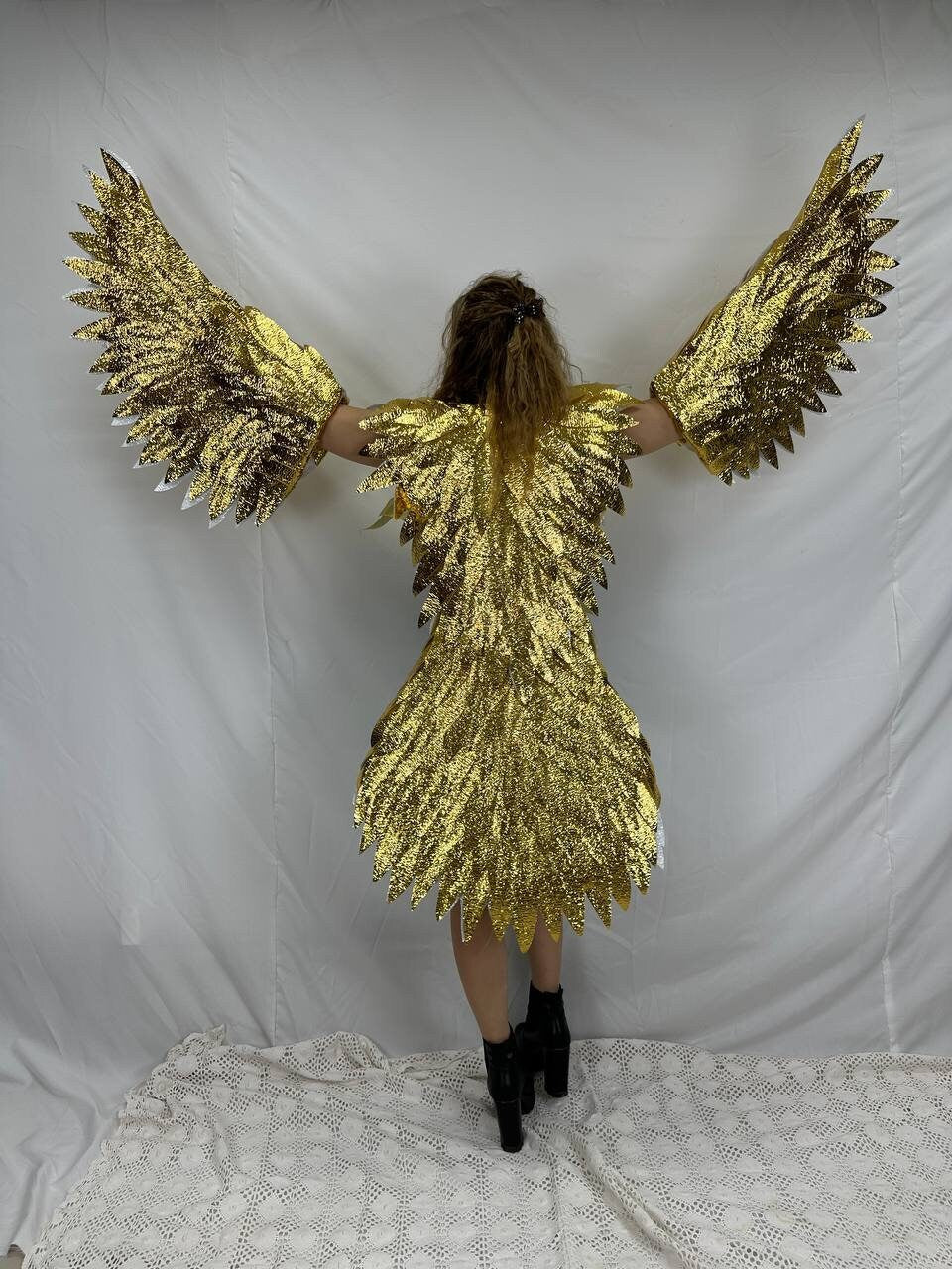May include: A gold sequined costume with large, outstretched wings. The costume appears to be a dress with attached arm and back pieces, creating a bird-like silhouette. The wings are detailed with individual feather shapes. The person is wearing black boots.