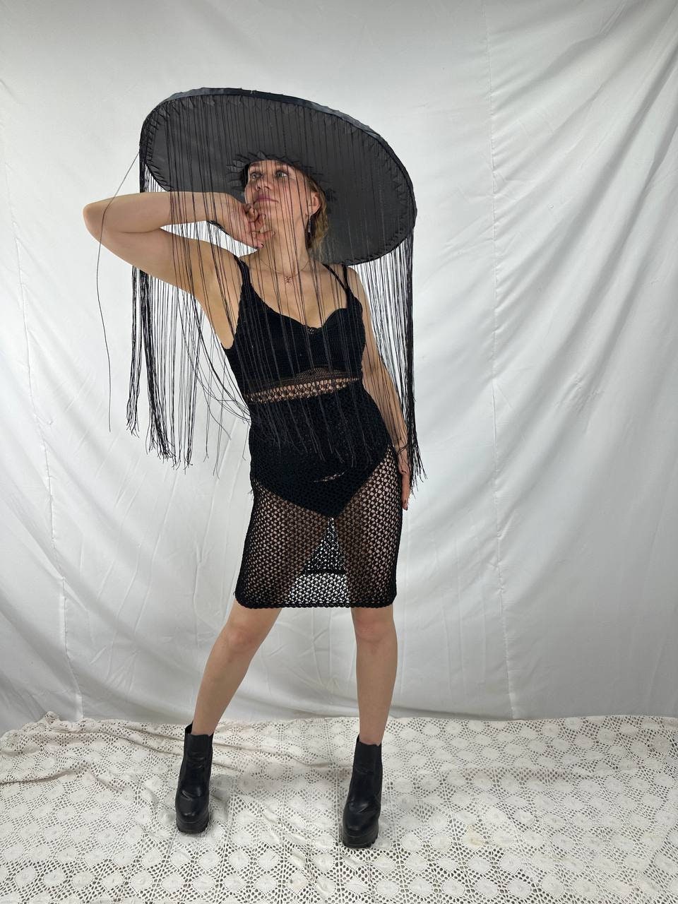 May include: A woman wearing a black crochet dress and a large black hat with fringe. The hat is wide brimmed and covers most of her face. She is wearing black boots.