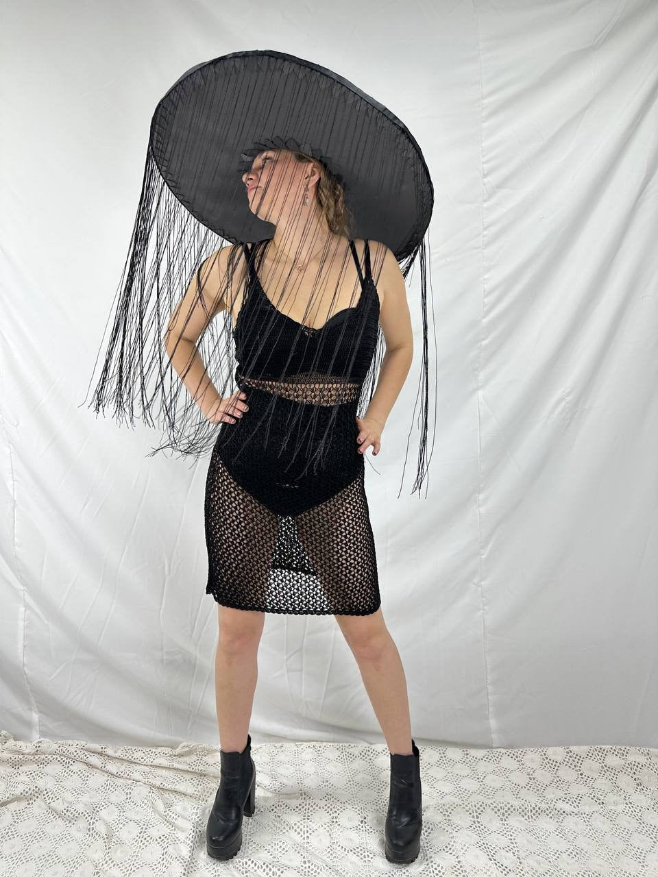 May include: A black crochet dress with a sheer design and a wide-brimmed black hat with fringe. The hat is large and covers most of the person's head.