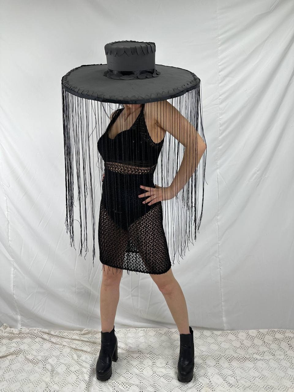 May include: A black wide-brimmed hat with a black ribbon and fringe hanging from the brim. The hat is worn by a person wearing a black mesh dress and black boots.