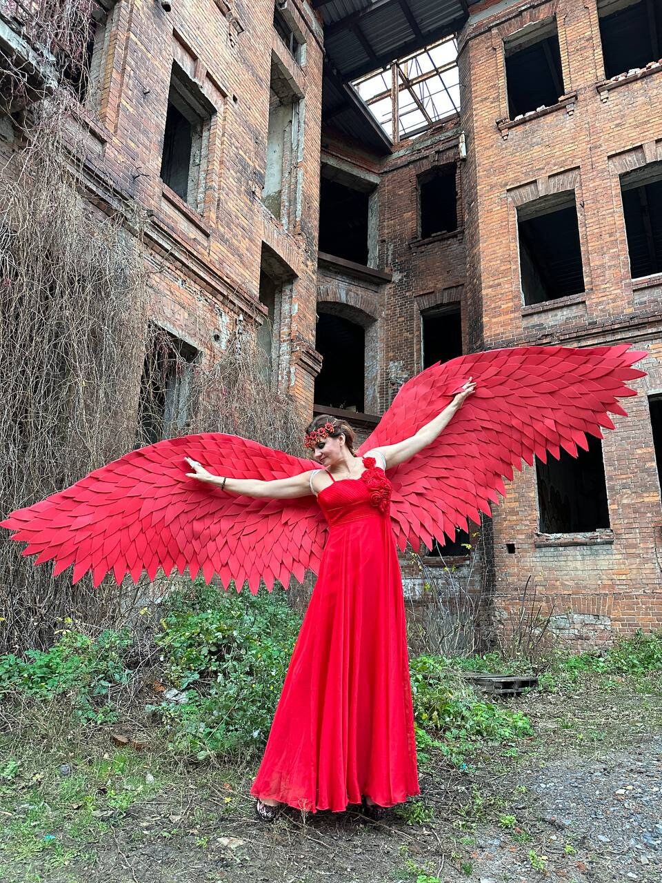 May include: A woman in a red dress with large red wings stands in front of a brick building with broken windows. The building appears to be abandoned.