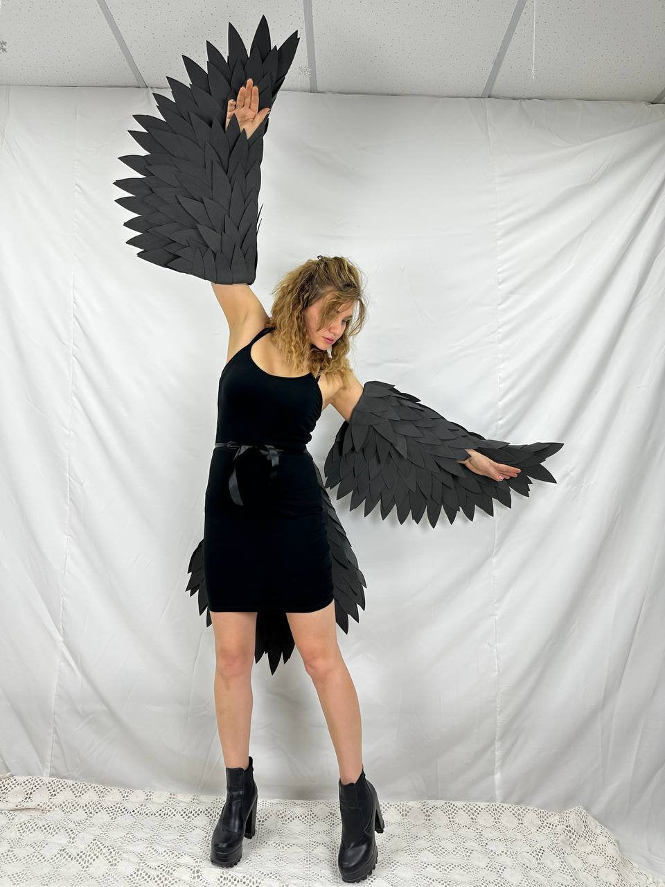 May include: A person wearing a black dress and black boots with large black wings made of feathers. The wings are attached to the arms and extend out to the sides.