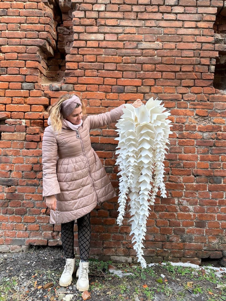 May include: A large, white, layered paper sculpture resembling a cascading floral arrangement. The artwork is held against a weathered brick wall, showcasing its intricate design and texture. The sculpture's form is reminiscent of a hanging plant.
