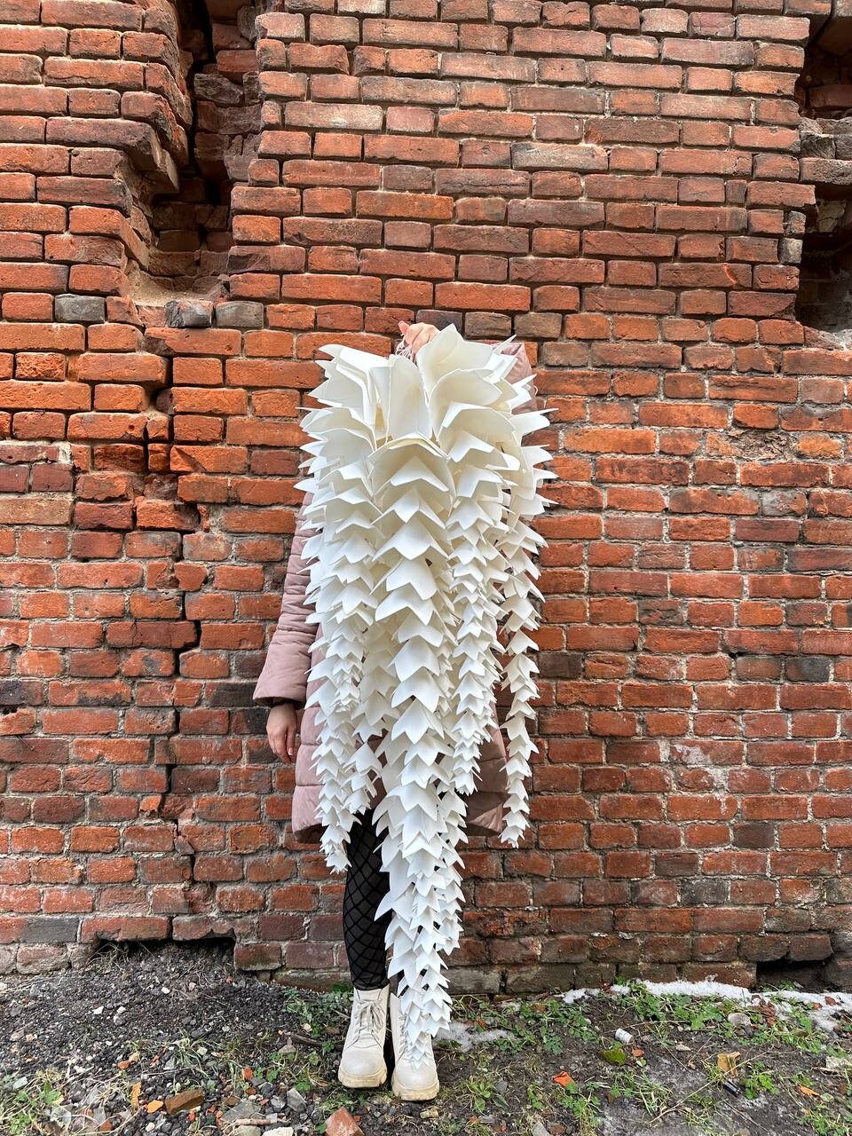 May include: A large, white, cascading paper sculpture with a layered, leaf-like design. The sculpture is held in front of a person standing against a weathered brick wall. The sculpture's shape resembles a hanging plant.