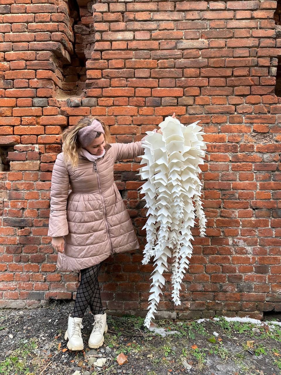 May include: A large, white, layered paper sculpture with a cascading, pointed design. The sculpture is held by a person in a pink coat, standing in front of a weathered brick wall. The sculpture's design resembles a stylized plant.