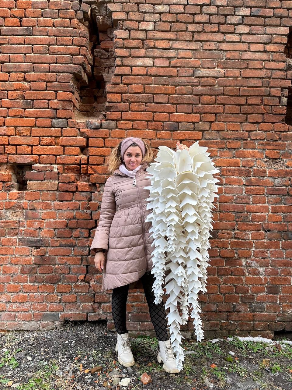 May include: A large, white, paper-like sculpture with a cascading, layered design. The artwork is held in front of a weathered brick wall. The sculpture's form resembles a stylized, elongated flower or plant.