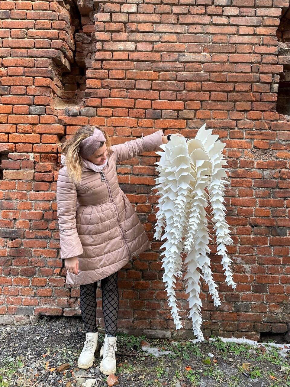 May include: A large, white, paper-like sculpture with a layered, cascading design. The sculpture is held in front of a weathered brick wall. The piece has a unique, organic shape, resembling a stylized plant or decorative element.