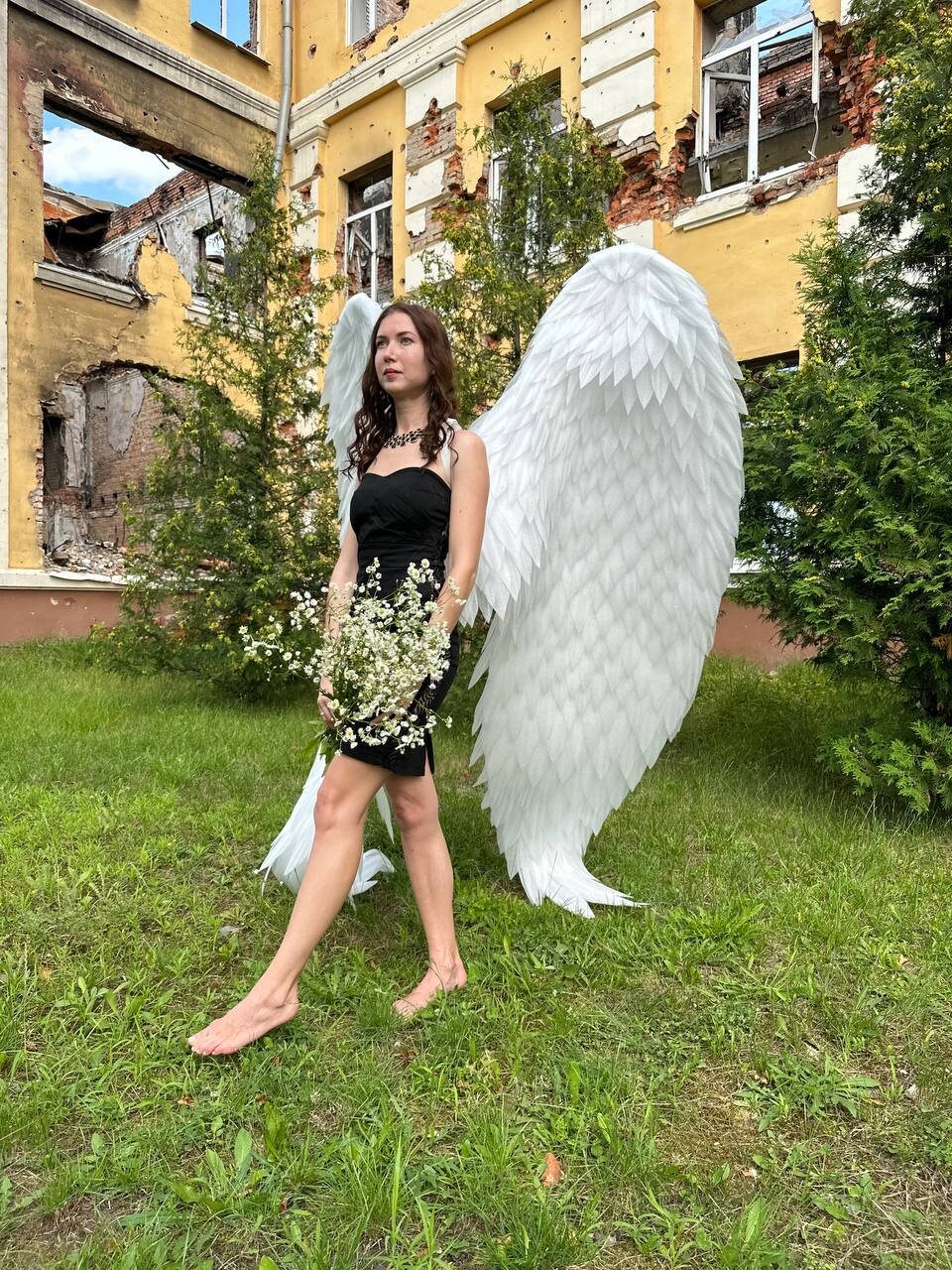 May include: A woman wearing a black dress and white angel wings stands in a grassy area in front of a damaged building. She is holding a bouquet of white flowers.