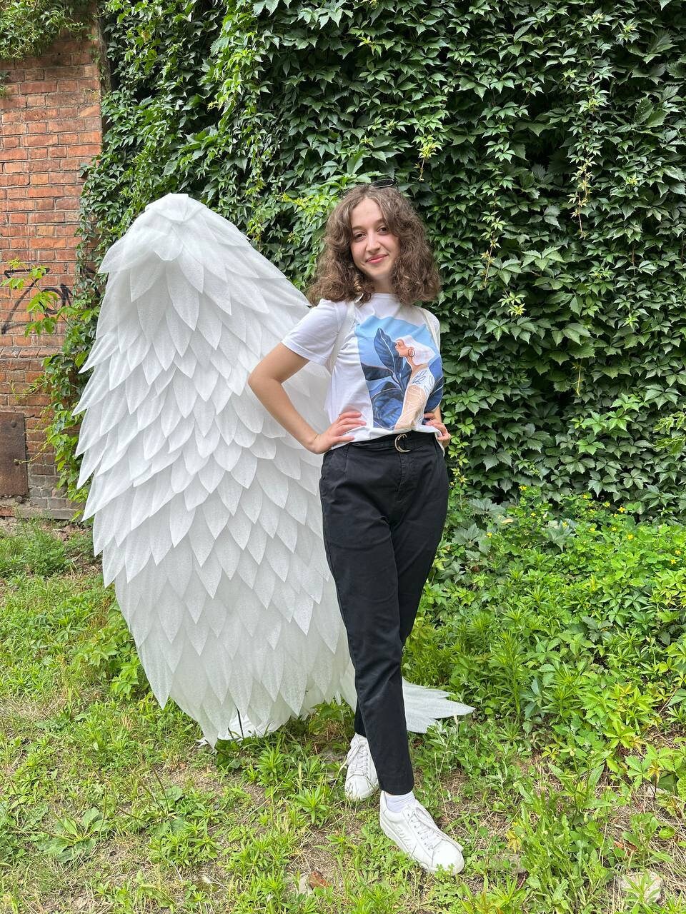 May include: A person wearing a white t-shirt with a graphic print and black pants stands in front of a large white feathered angel wing prop.