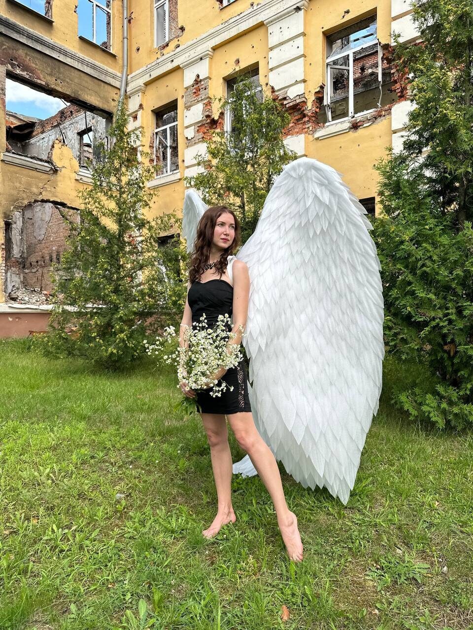 May include: A woman wearing a black dress and white angel wings stands in a grassy area in front of a building with bullet holes. She is holding a bouquet of white flowers.