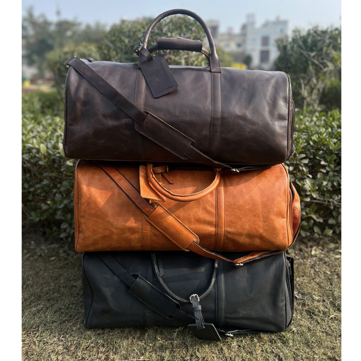May include: Three leather duffel bags stacked on top of each other. The top bag is brown, the middle bag is orange, and the bottom bag is black.