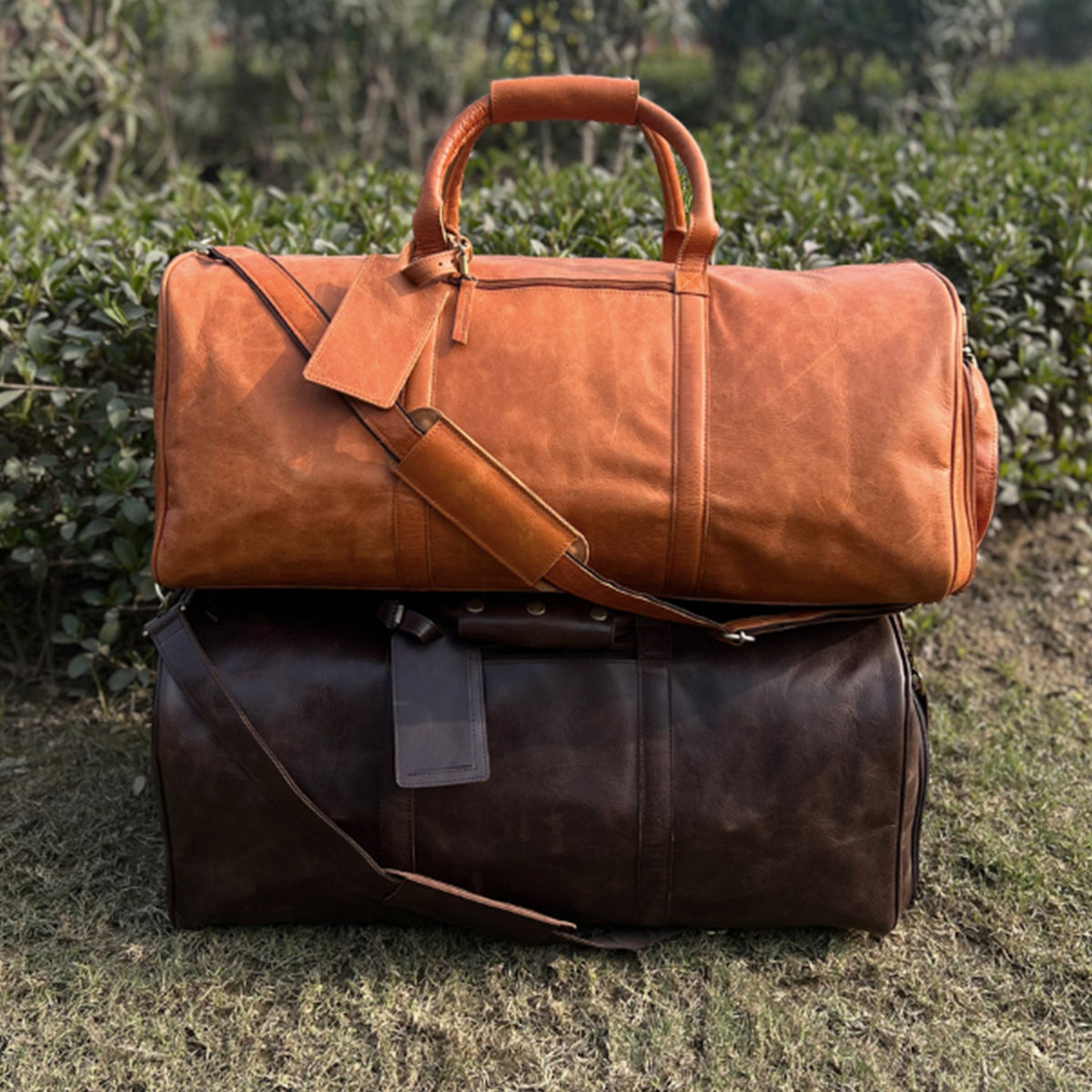 May include: Two leather duffel bags stacked on top of each other. The top bag is a light brown color and the bottom bag is a dark brown color.